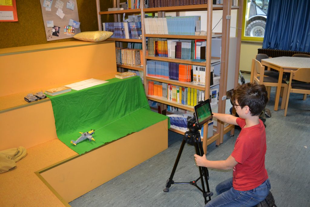 Junge kniet vor einem Tablet, das in ein Stativ eingeklemmt ist. er filmt ein Spielzeug-Flugzeug, das auf einem grünen Hintergrund liegt.