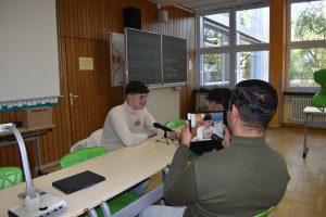 Drei Jugendliche sitzen sich an Schultischen gegenüber. Einer filmt mit dem Tablet, einer spricht in ein Mikrofon, das auf dem Tisch steht, einer sitzt ihm gegenüber.