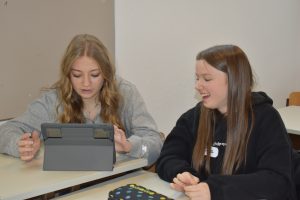 zwei Jugendliche sitzen an einem Schultisch, ein aufgestelltes Tablet vor ihnen. Eine schaut auf den Bildschirm und tippt darauf, die andere schaut auf den Bildschirm und lacht.