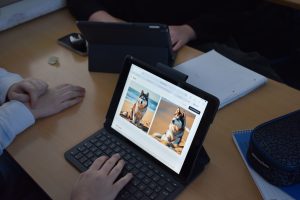 Ein Schreibtisch ist zu sehen, auf dem ein Tablet steht, auf dem Bildschirm sind Fotos eines Hundes zu sehen, wobei es sich um KI generierte Bilder handelt. Auch die Hände von drei Personen sind zu sehen, Ein Block, ein Handy und ein weiteres Tablet, dessen Bildschirm weggedreht ist.