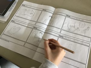auf einem A3 Blatt wurde ein Storyboard gezeichnet. Das Papier liegt auf dem Tisch, darauf die Hand einer jugendlichen Person mit Bleistift in der Hand. Zu sehen sind einzelne Szenen des geplanten Videos.