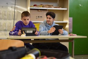 zwei jugendliche Jungs sitzen an einem Schultisch und haben Tablets in der Hand. Einer schaut auf den Bildschirm, einer zur Seite.