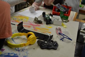 Tisch mit einer fleckigen Abdeckfolie und Elektroschrott Teilen, einer Schutzbrille und Hände von einem Kind mit einem Handschuh.