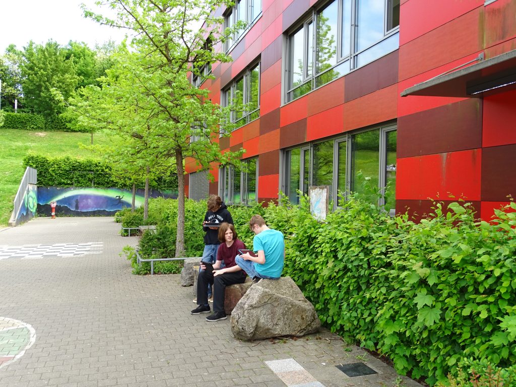 drei Jugendliche sitzen und stehen vor einem Schulgebäude und arbeiten an Tablets. Das Gebäude ist rot, es ist viel Grün durch Büsche und Wiese zu sehen.