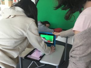 Jugendliche liegt mit dem Kopf auf einem Schultisch, neben ihr ein roter Hut, dahinter ein Greenscreen. Sie wird von zwei Mitschülerinnen mit einem Tablet gefilmt.