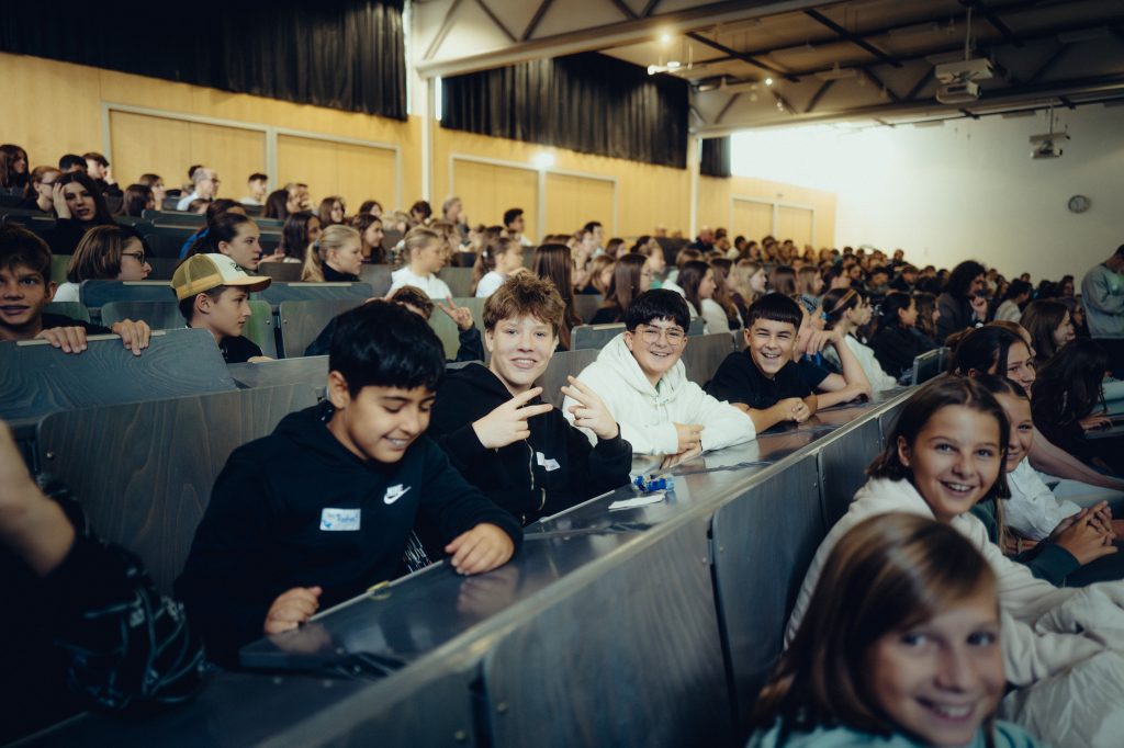 ein großer Hösal voller junger Schüler*innen. Im Fokus drei lachende Jungen.