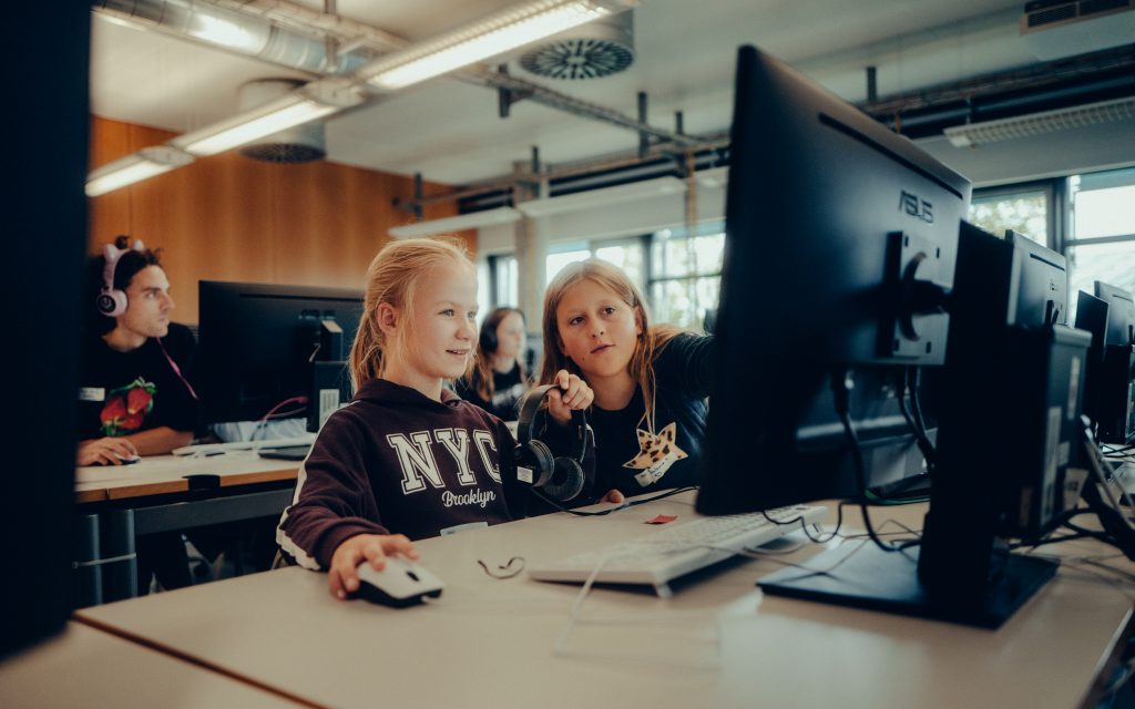 zwei schülerinnen schauen begeistert auf ihren computerbildschirm. im hintergund hilft der workshopleiter und trägt dabei einen rosa kopfhörer mit katzenohen.
