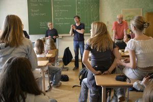 ein volles Klassenzimmer. vor der tafel steht der Workshop Leiter und spricht. an der tafel sieht man eine Tabelle mit Projekten und Arbeitsschritten samt vieler Erledigt-Haken.