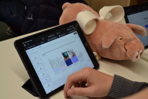 Ein aufgestelltes Tablet und die Hand einer Person ist zu sehen, daneben auf dem Tisch liegt ein Stoffschwein.