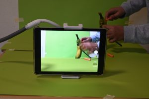 Auf einem Tabletbildschirm ist eine Banane zu sehen, die jemand im Hintergrund hält. Die Banane hat ein aufgemaltes Gesicht und steht vor einem grünen Hintergrund.