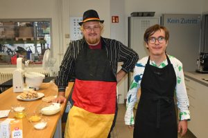 Zwei Teilnehmende schauen freundlich in die Kamera. Sie tragen Schürzen und neben ihnen ist eine Küchenarbeitsfläche. Auf ihr stehen Zutaten in Schüsseln und eine Rührmaschine.