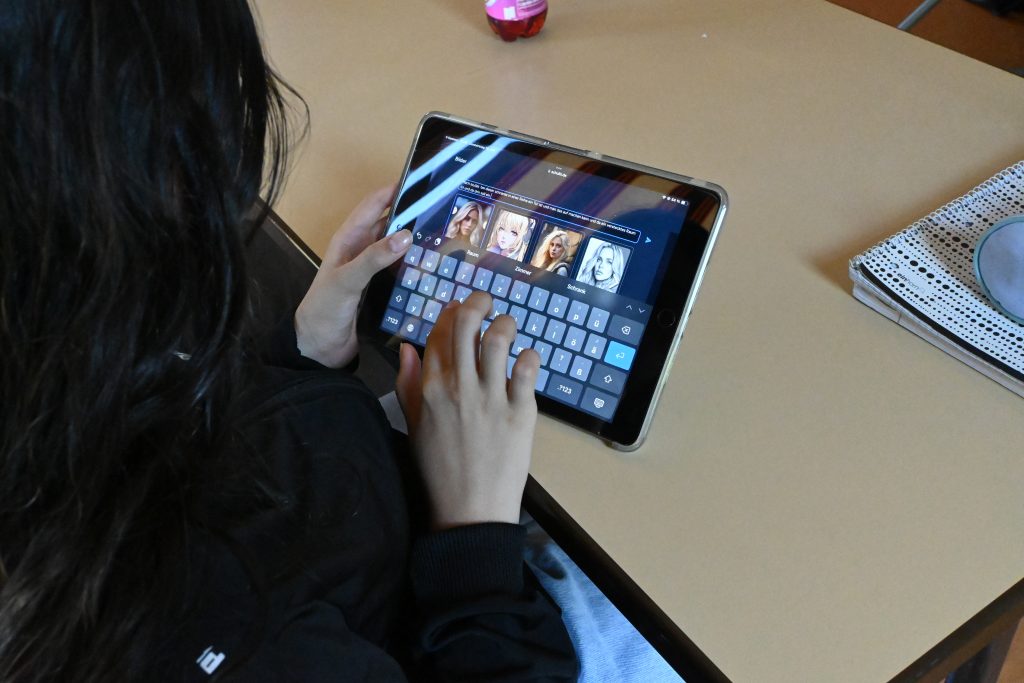 Eine Jugendliche sitzt am Schultisch und hat ein Tablet in der Hand, auf dem sie etwas tippt.