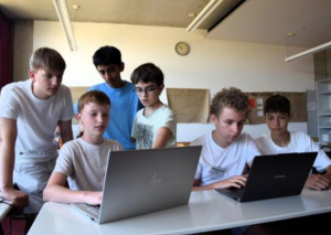 zwei Schüler arbeiten konzentriert an Laptops. Um sie herum stehen und sitzen vier weitere die gespannt auf die Monitore schauen.
