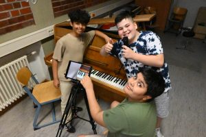 drei Kinder posieren mit Tablet und Mikrofon vor einem Klavier, sie blicken in die Kamera.