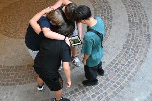 insgesamt vier Jugendliche stehen im Kreis um ein Tablet und blicken darauf, teilweise legen sie die Arme umeinander. Das Foto wurde von oben aufgenommen.