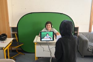 Ein Kind sitzt an einem Tisch, ein Greenscreen im Hintergrund, mit Kopfhörern augesetzt, er lacht freundlich in die Kamera eines ipads. Der Bildschirm des ipads ist zu sehen, daneben ein weiteres Kind.