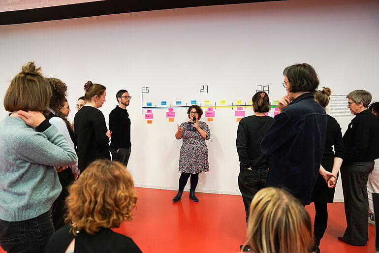 Moderatorin hät ein Mikrofon in der Hand und steht vor einer Wand mit einem Zeitstrahl. Drumherum sind die Botschafter*innen vom Kurswechsel