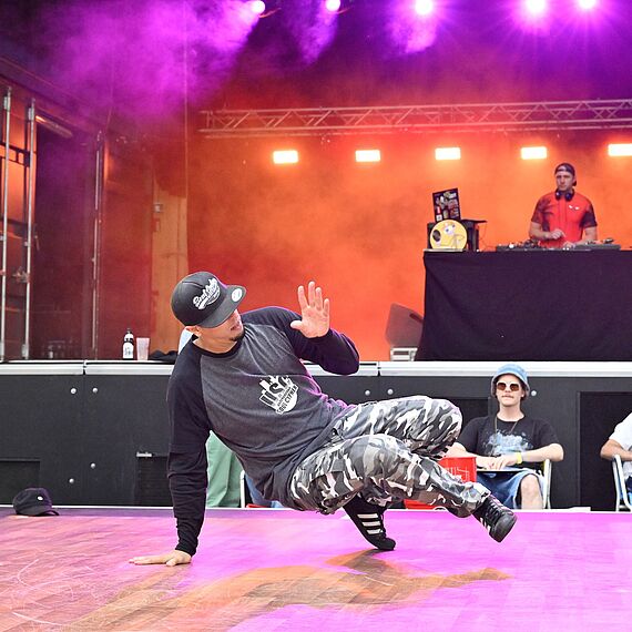 eine Person macht break dance vor einer Bühne auf det ein DJ an einem DJ Pult steht. Die Bühne ist orange und lila beleuchtet