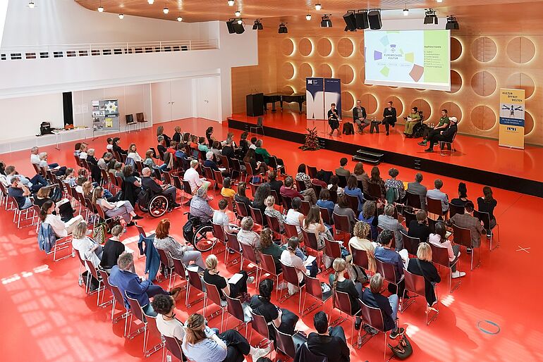 Publikum ca. 100 Personen von oben fotografiert, auf der Buehne sitzen fünf Personen