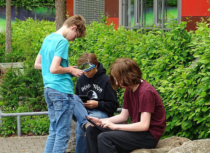 Drei Jungs sitzen und stehen draußen und schauen auf ihre Ipads runter.