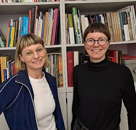 Vor einem Bücherregal stehen zwei Frauen und lächeln in die Kamere. sie haben beide eine Brille.