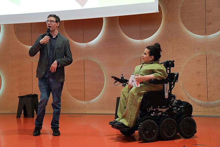 Mann spricht auf der Buehne ins Mikro und daneben sitzt eine Frau im Rollstuhl