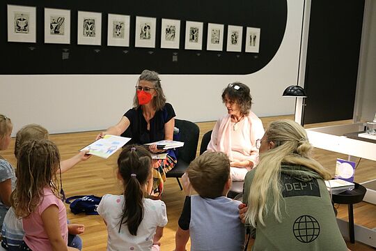 Antje Meyer mit roter Maske sitzt vor Kindern, die mit dem Rücken zum Betrachter sitzen. 