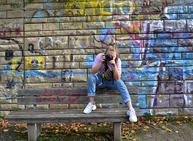 Mädchen sitzt auf der Lehne einer Parkbank und schaut durch eine Kamera.