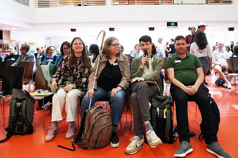 Frau mit Langstock sitzt zwischen zwei Maennern und einer Frau in einer Stuhlreihe