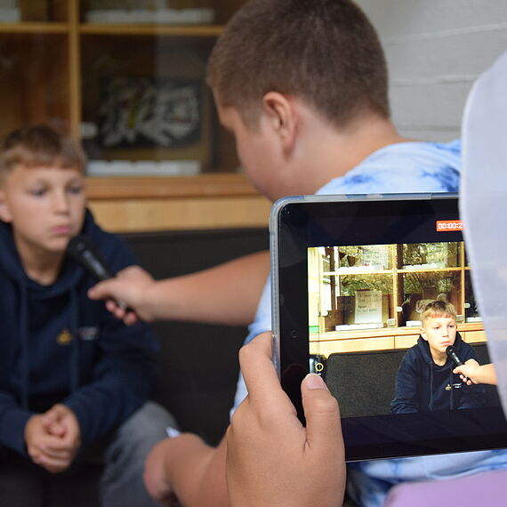 Zwei Jungs interviewen sich mit Mikrofon und werden dabei von einem Jungen mit Tablet gefilmt. 