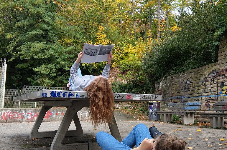 Ein Mädchen fotografiert ein auf einer Tischtennisplatte liegendes Mädchen mit Zeitschrift in der Hand.
