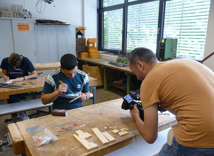 Jugendlicher sitzt am Tisch in der Werkstatt und arbeitet mit Holz. Dabei wird er von einem Mann gefilmt.