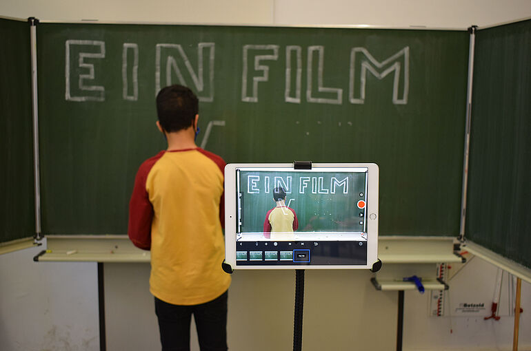 Junge steht mit Rücken zur Tafel und schreibt, wird dabei mit Tablet gefilmt
