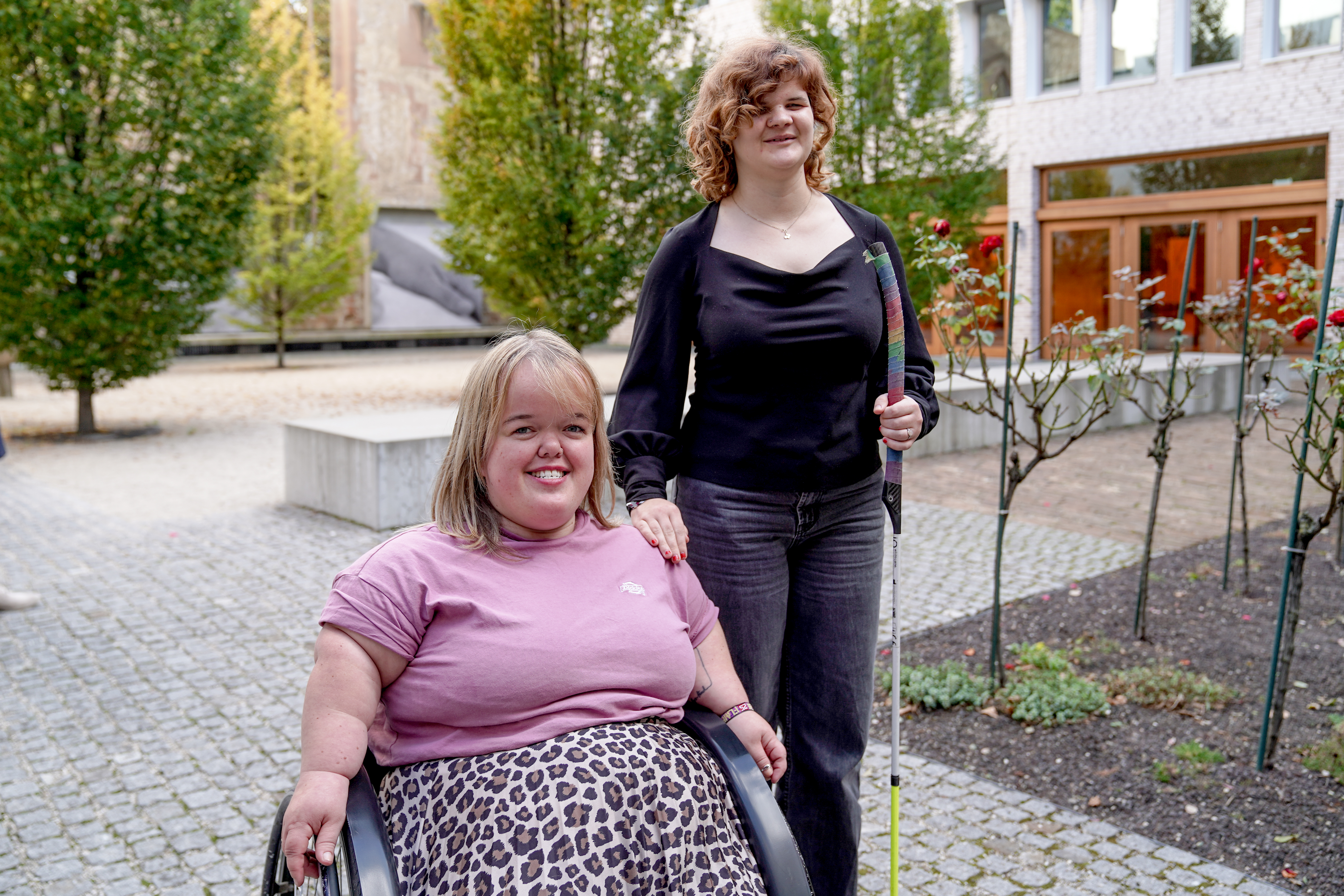 Auf dem Bild sieht man zwei Frauen, links eine Frau im Rollstuhl ,lächelnd, rechts von ihr eine Frau mit Blindenstock.